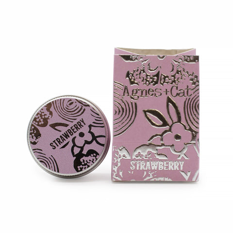 Strawberry lip balm tin beside its pink printed carton, shown close up against a white background