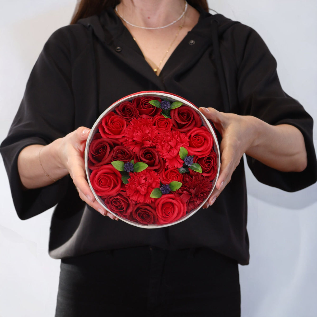 A person holds a round box filled with red roses and small red flowers.