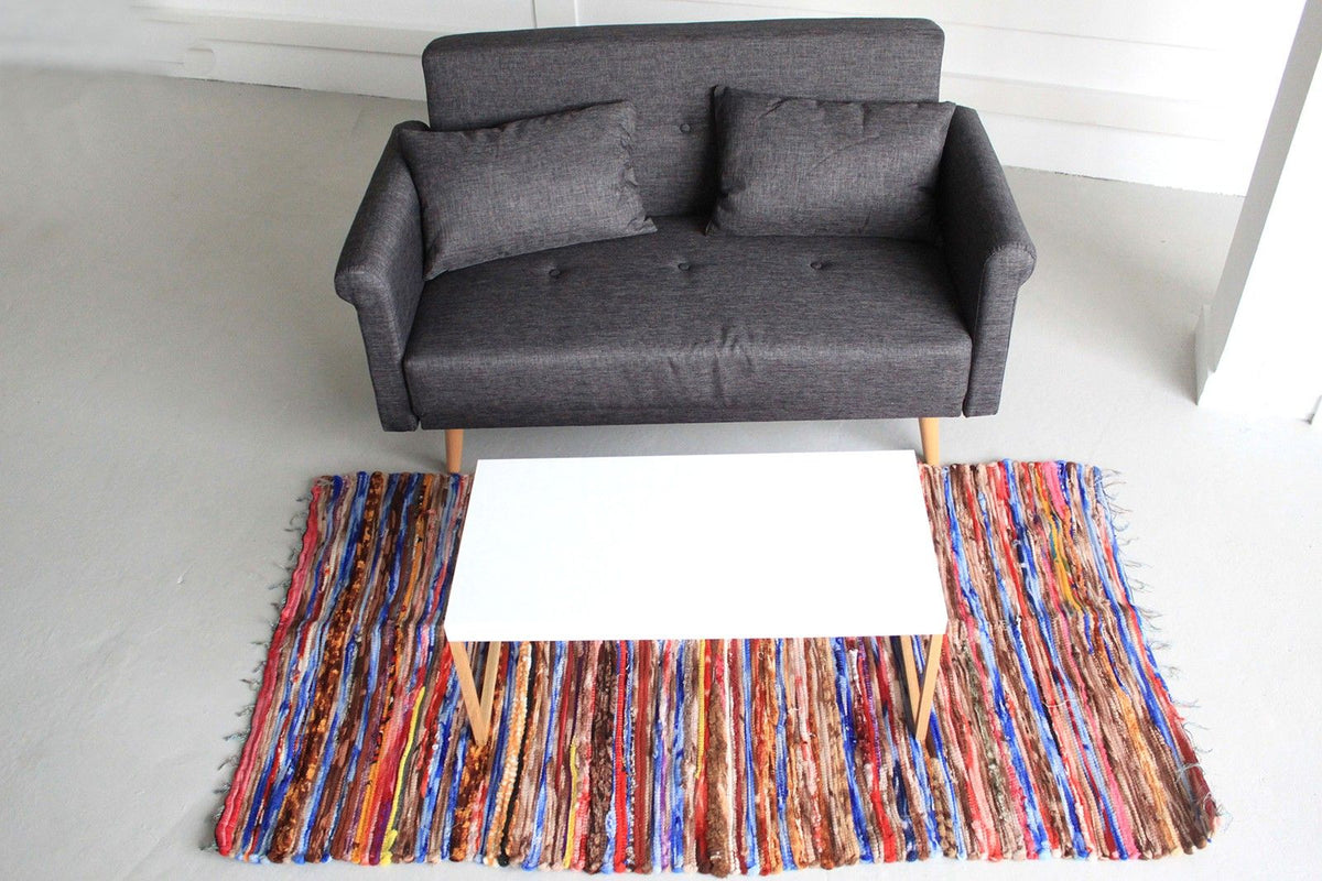 A multicoloured striped rug on a light floor beneath a small white table and beside a grey sofa.