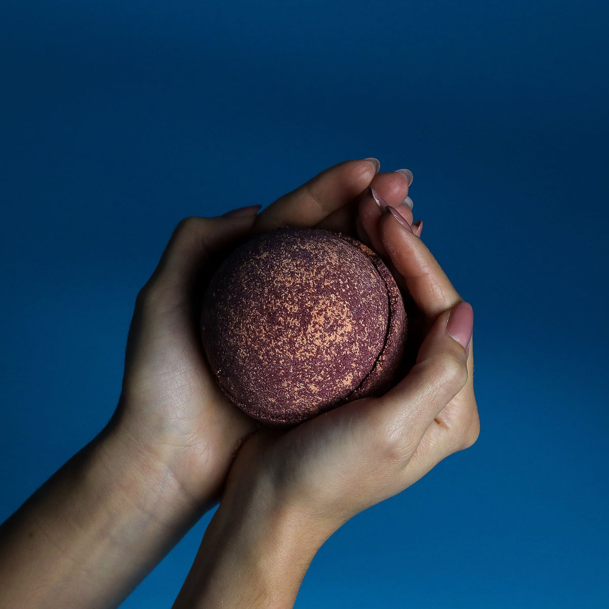 Two hands holding a round brown bath bomb against a blue background.
