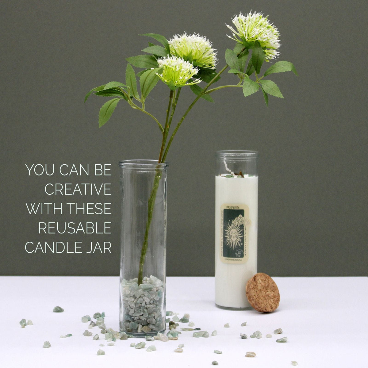 A white candle in a glass jar beside a tall glass vase with flowers and scattered stones.