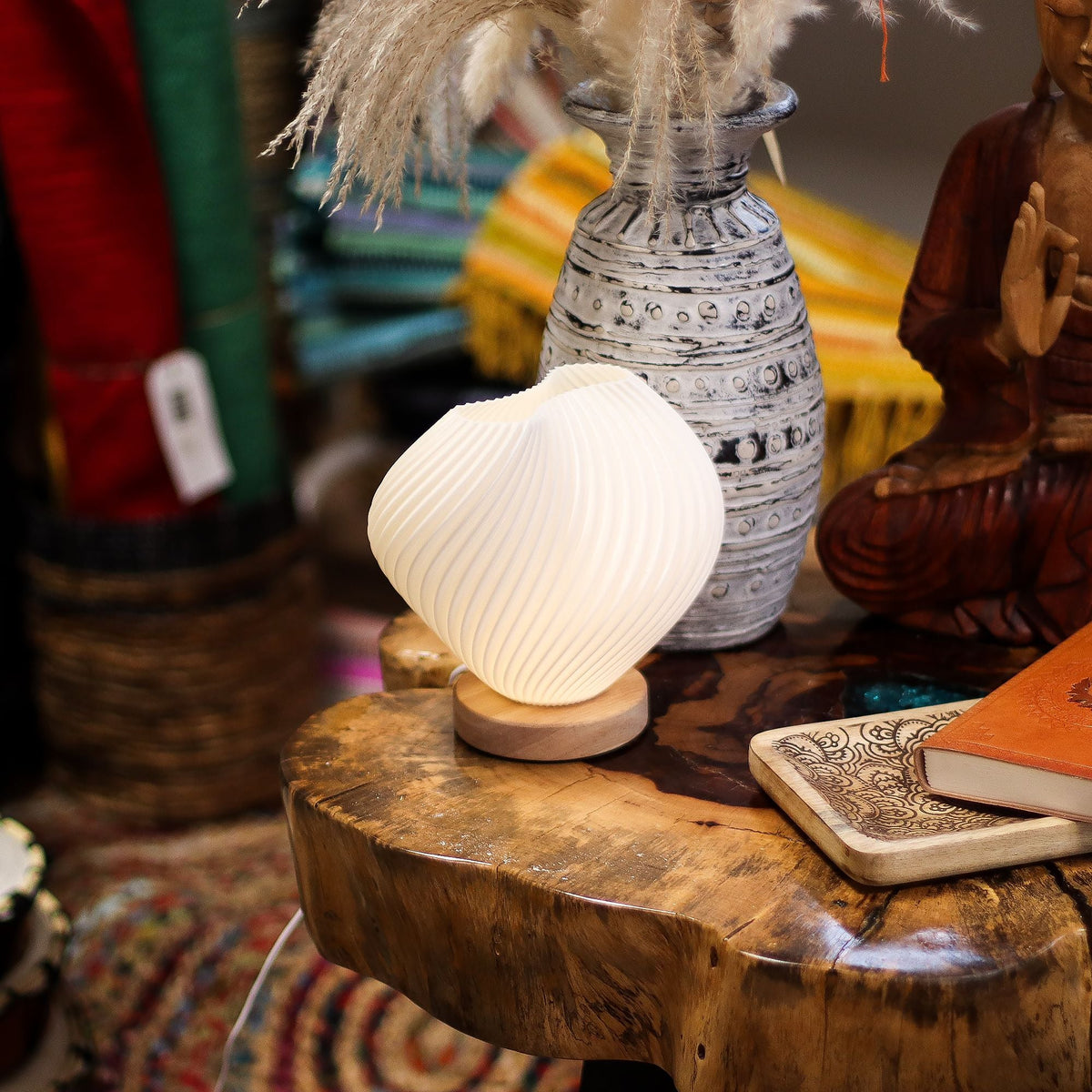 Small white shell-shaped lamp on a round wooden base on a carved wooden table, with ornaments and a notebook nearby.