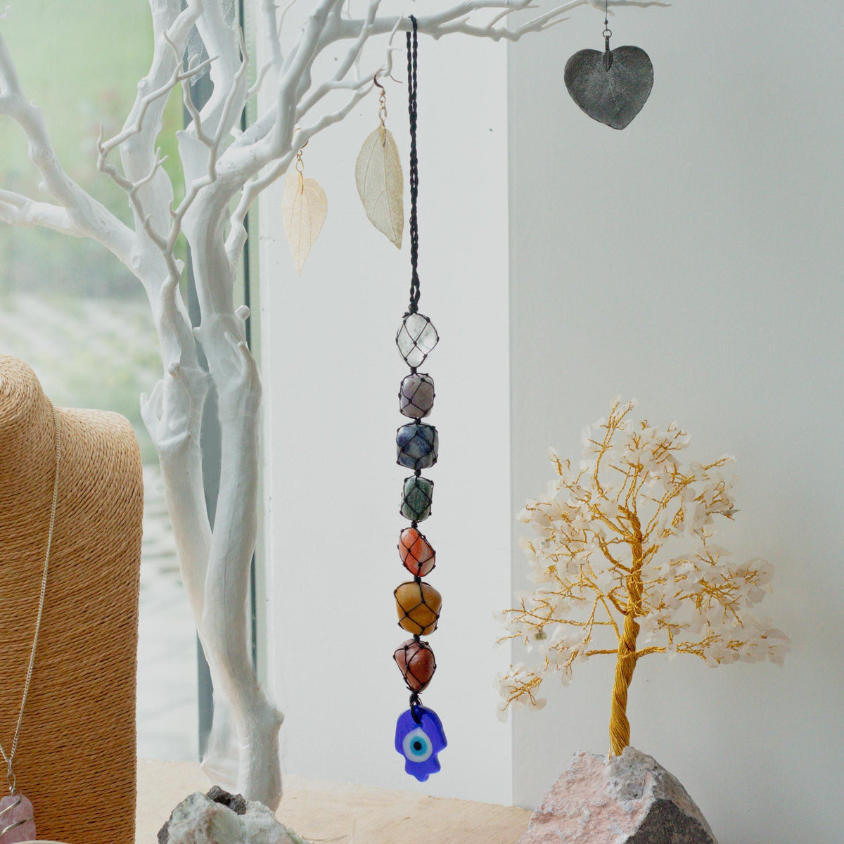 Hanging chakra stone pendant with coloured stones and a blue charm beside decorative tree branches.