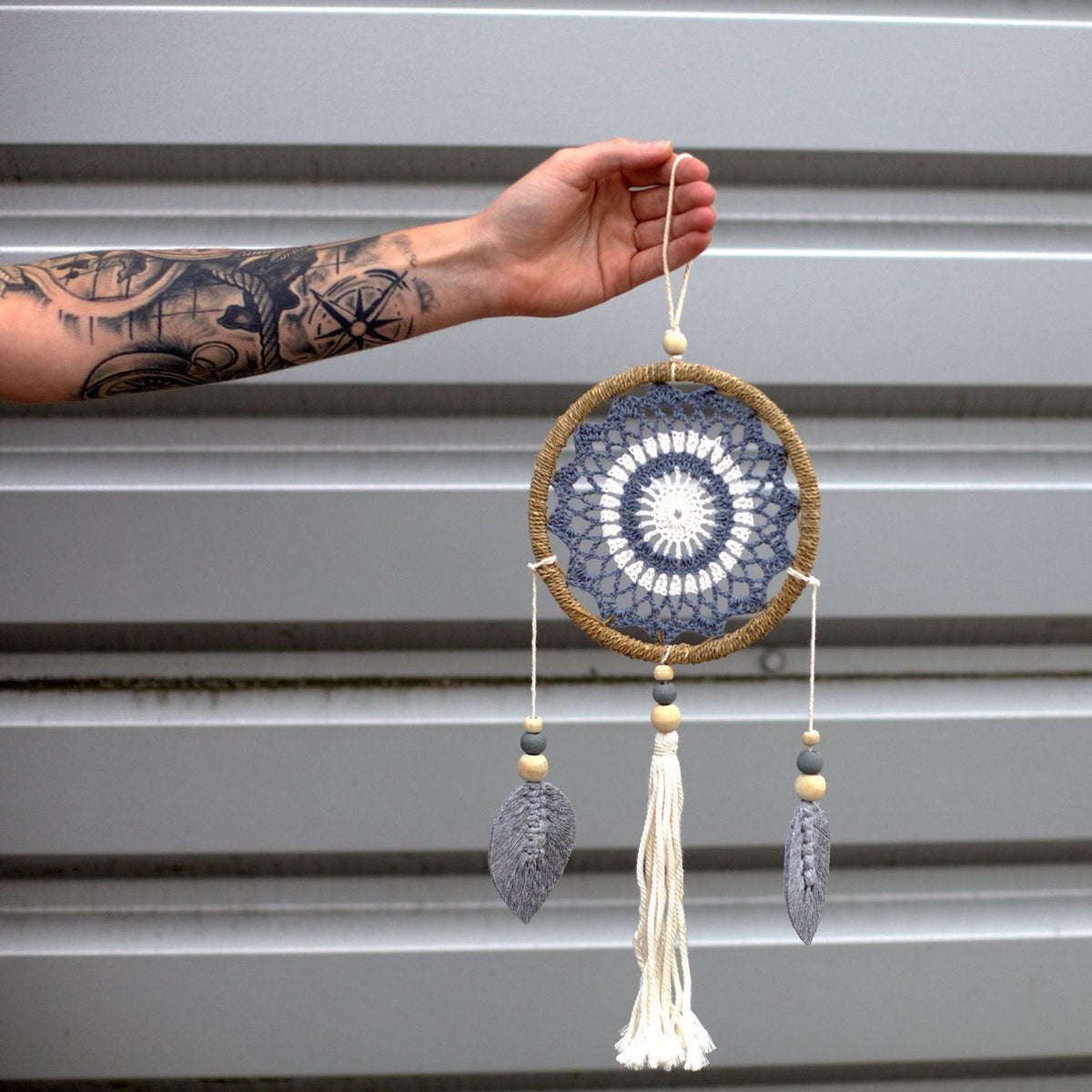 A hand holding a grey and cream dreamcatcher with three hanging tassels.