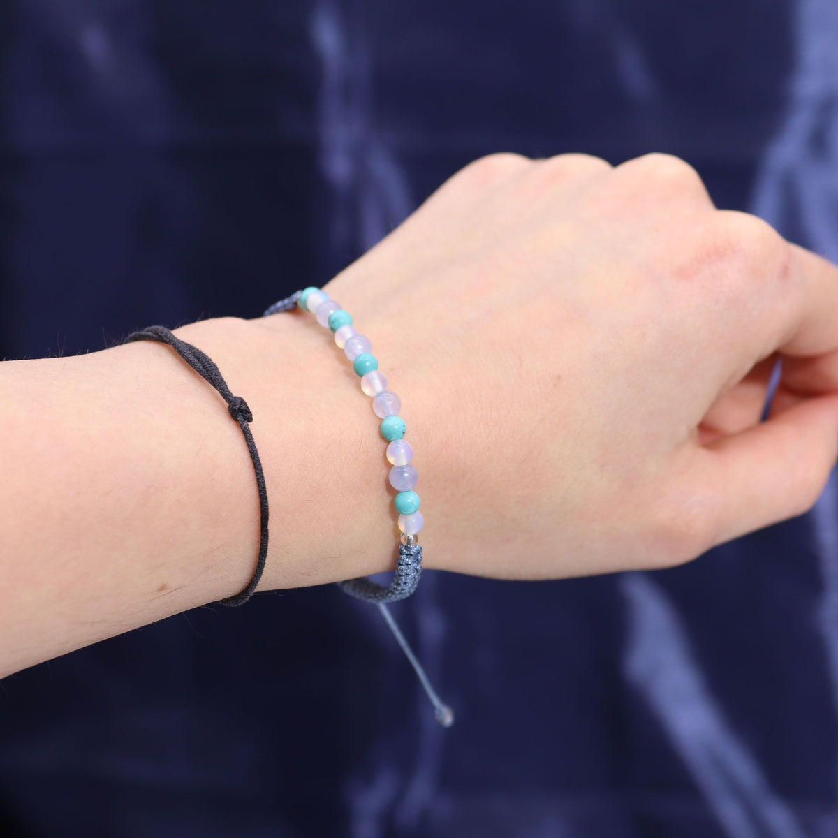 A hand wearing a beaded bracelet and a black cord bracelet against a dark blue background.