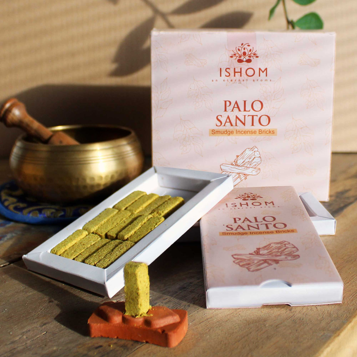 A terracotta incense burner, a brass bowl with spoon, and two boxes of Palo Santo incense bricks on a wooden table.