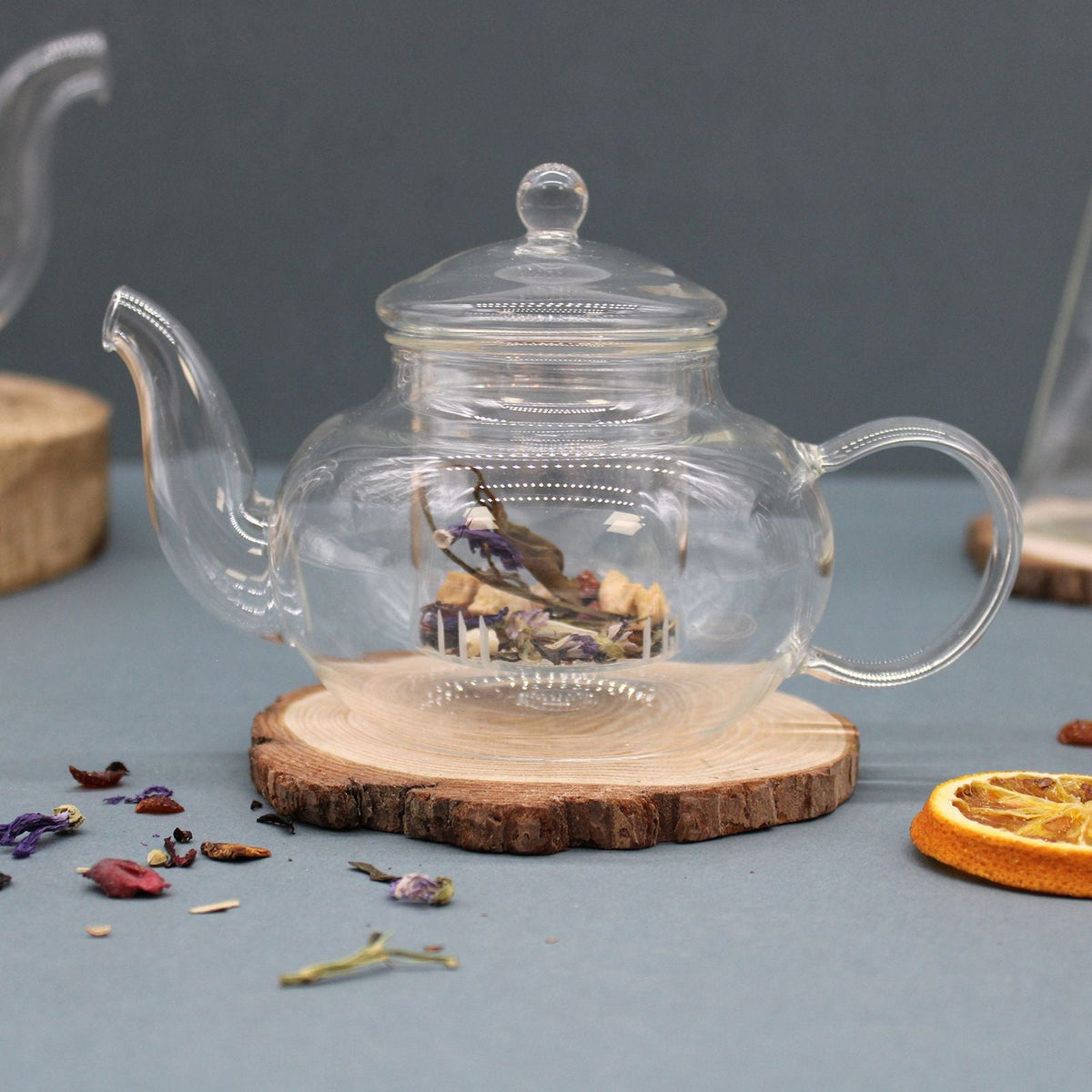 Clear glass teapot with a lid on a round wooden slice, surrounded by dried flowers and fruit.