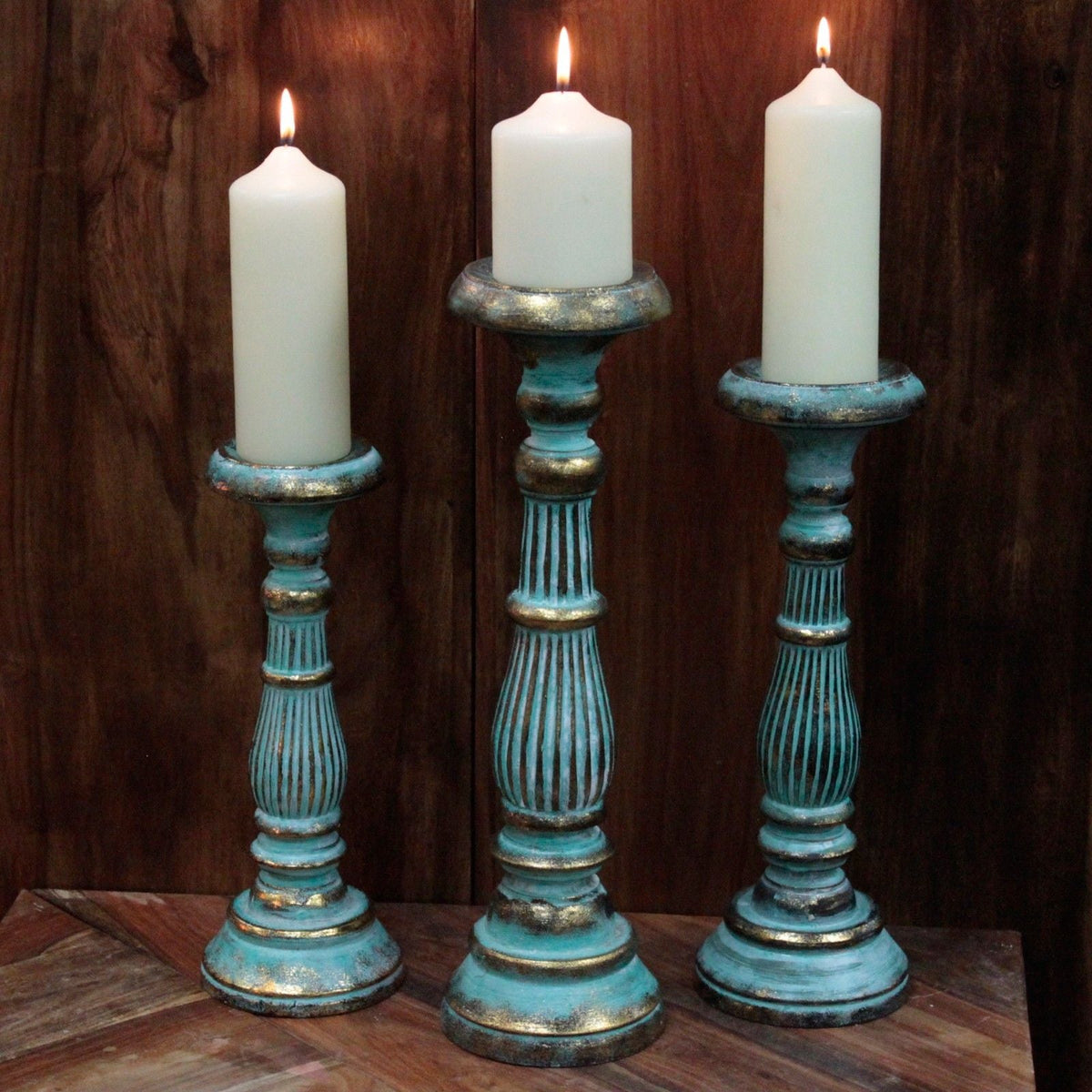 Three graduated wooden candlesticks in a distressed turquoise-and-gold finish, each holding a lit white pillar candle, on a wooden table against a dark wood-panelled background.