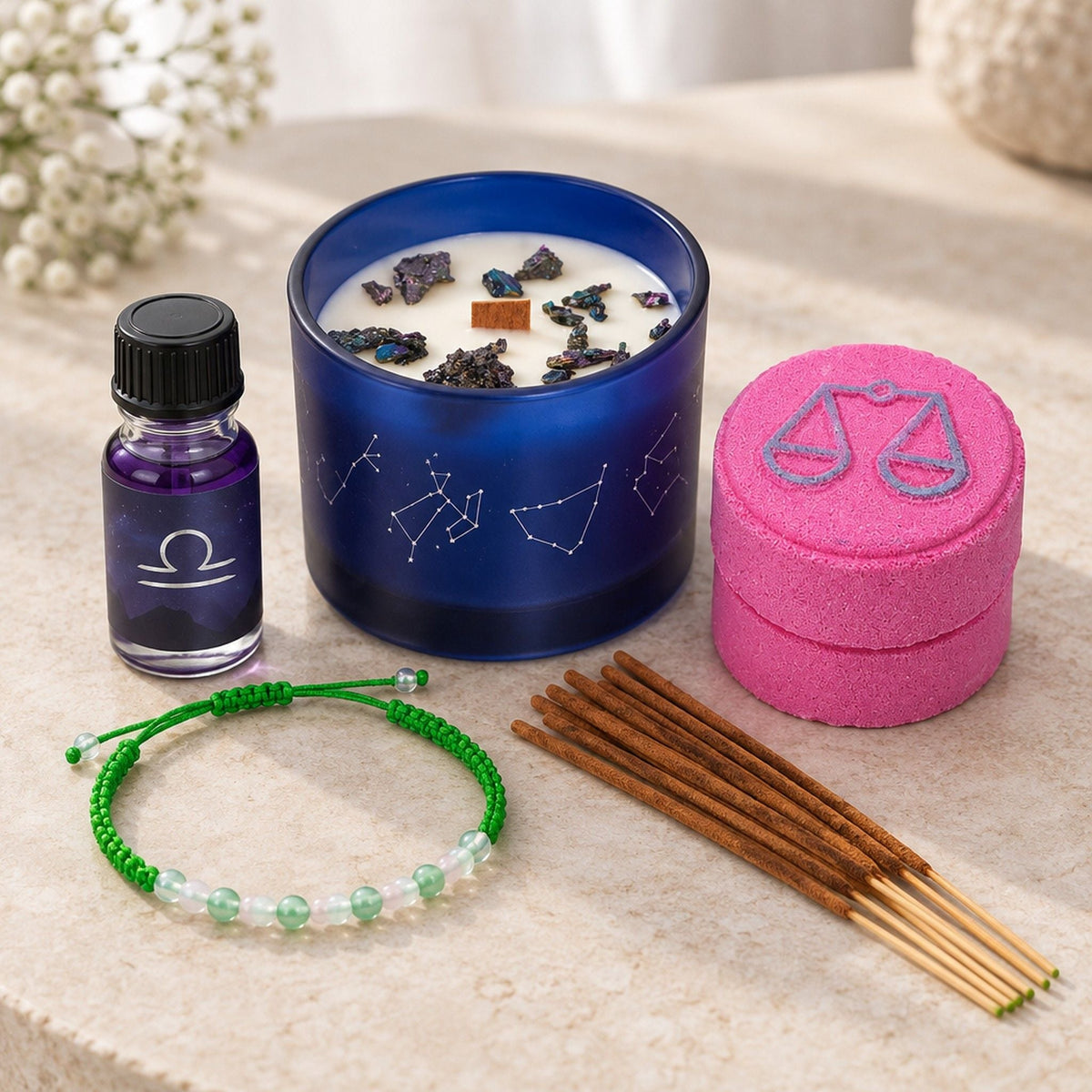A blue candle, purple oil bottle, pink bath fizzer pot, green beaded bracelet and incense sticks arranged on a stone surface.
