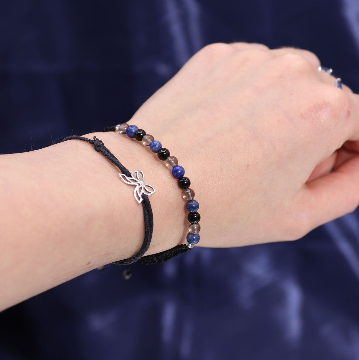 A hand wearing two bracelets, one beaded and one black cord with a small silver butterfly charm.