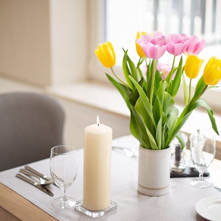 A lit ivory pillar candle on a dining table beside glasses and a vase of tulips.