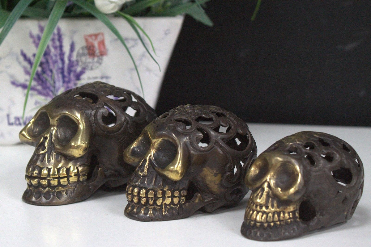 Three handmade brass skulls with openwork detail and polished highlights lined up on a white shelf, with a potted plant behind them