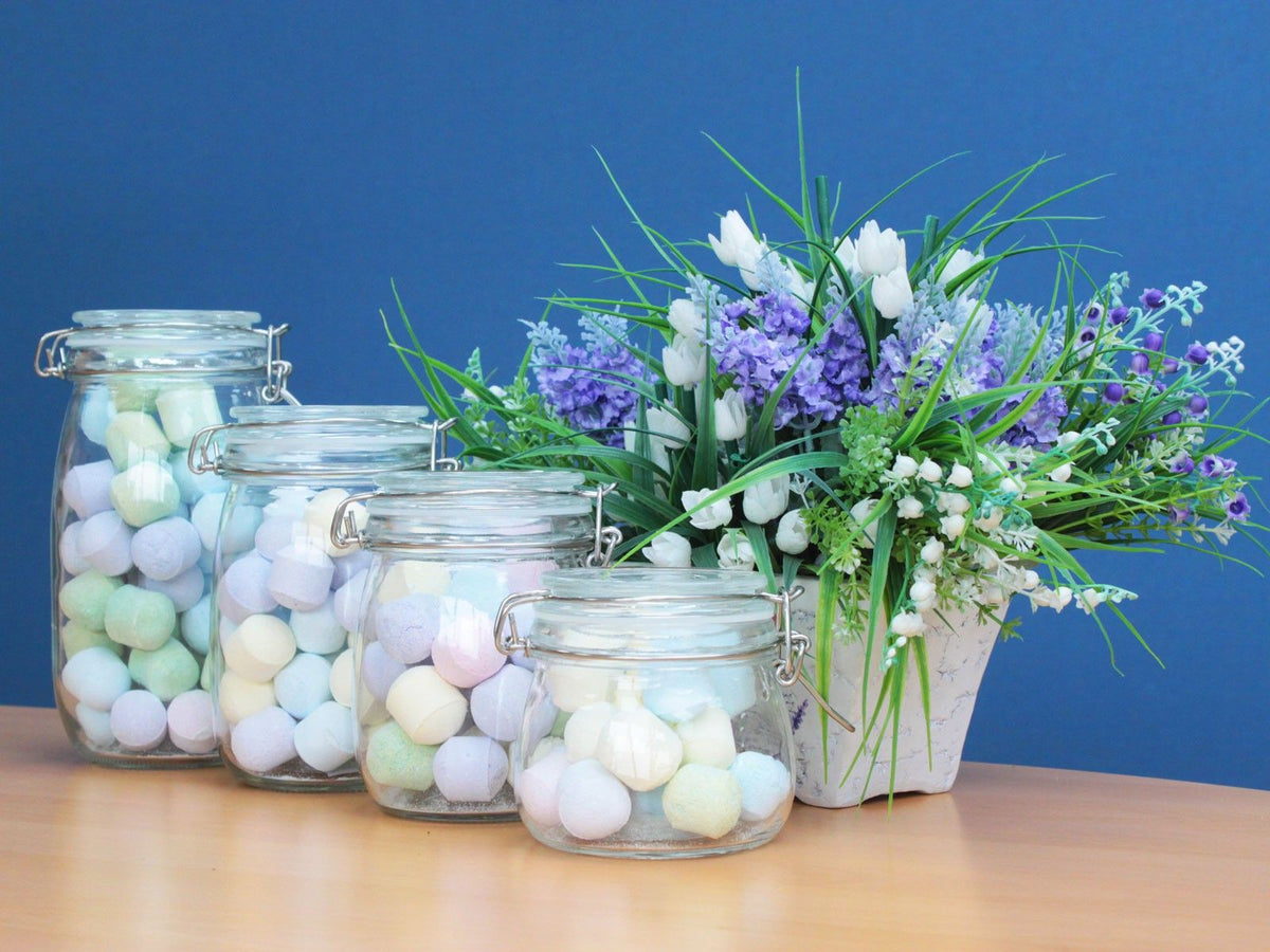 Four glass clip-top jars filled with pastel balls on a wooden surface beside purple and white flowers.