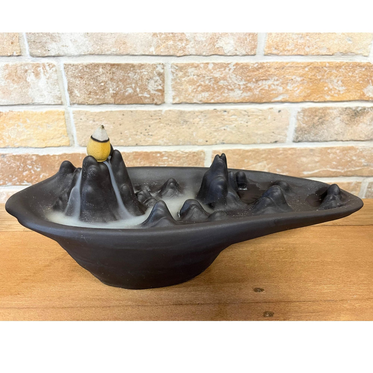 Mountain Scape backflow incense burner on a wooden table against a brick wall, with a lit cone on its peak and smoke flowing down the slopes