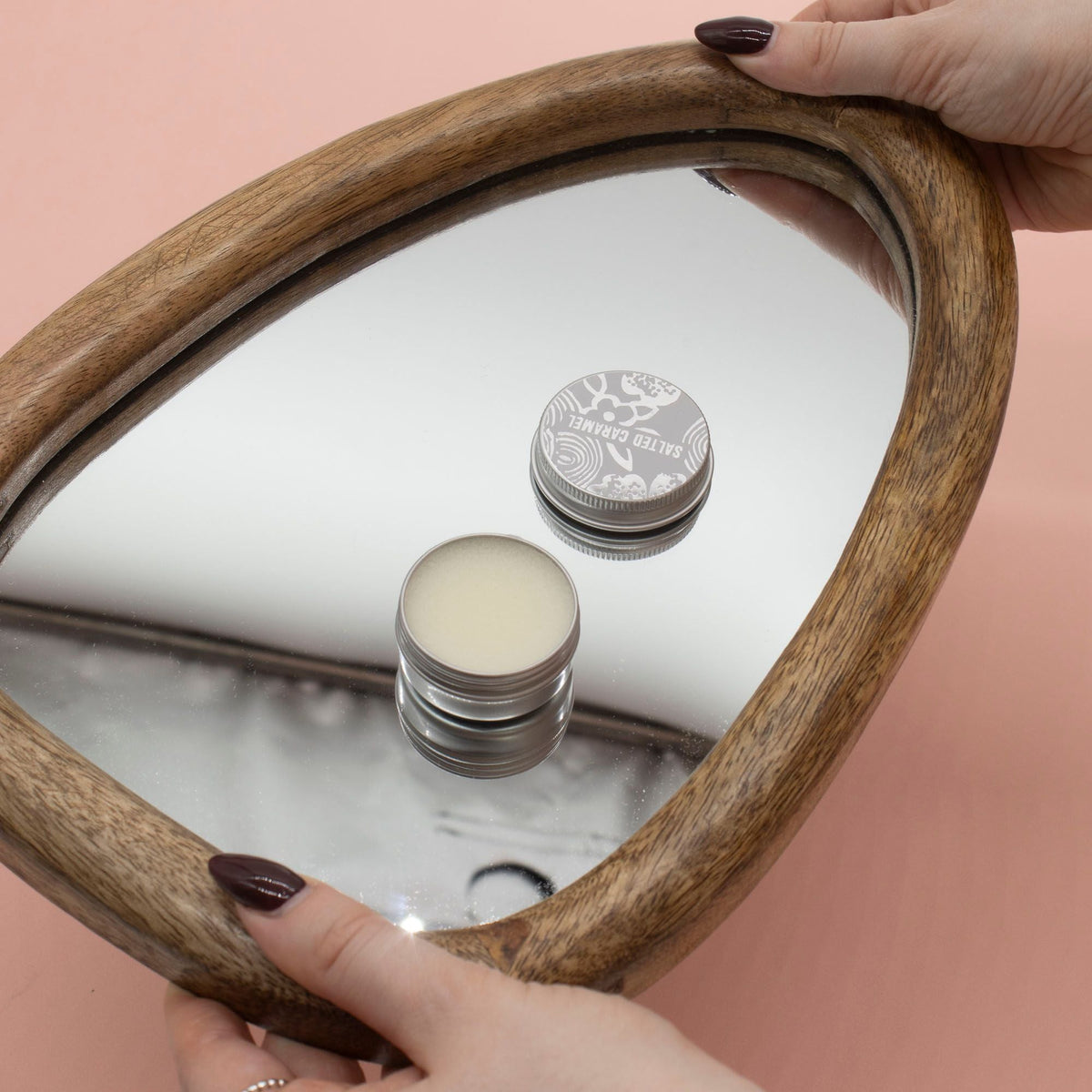 An open silver lip balm tin and its lid on a mirrored wooden tray held by two hands.