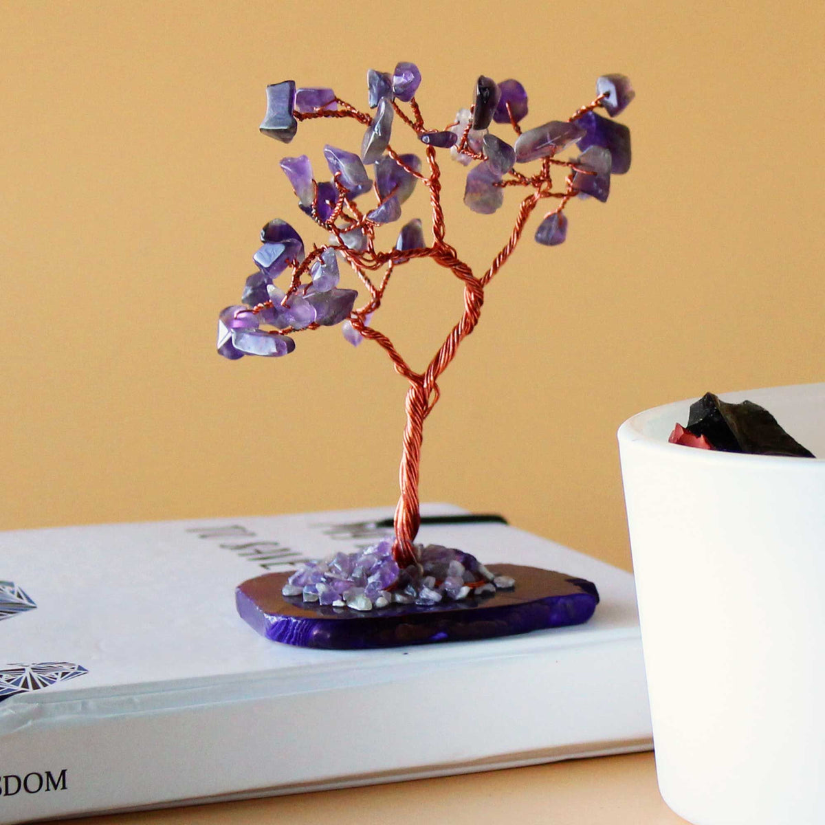 A small amethyst gemstone tree on a purple agate base beside a white box and a cup.