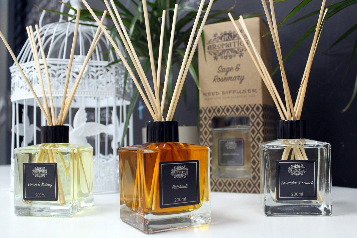 Three square reed diffusers with sticks on a white surface, with a decorative lantern and a boxed diffuser behind them.
