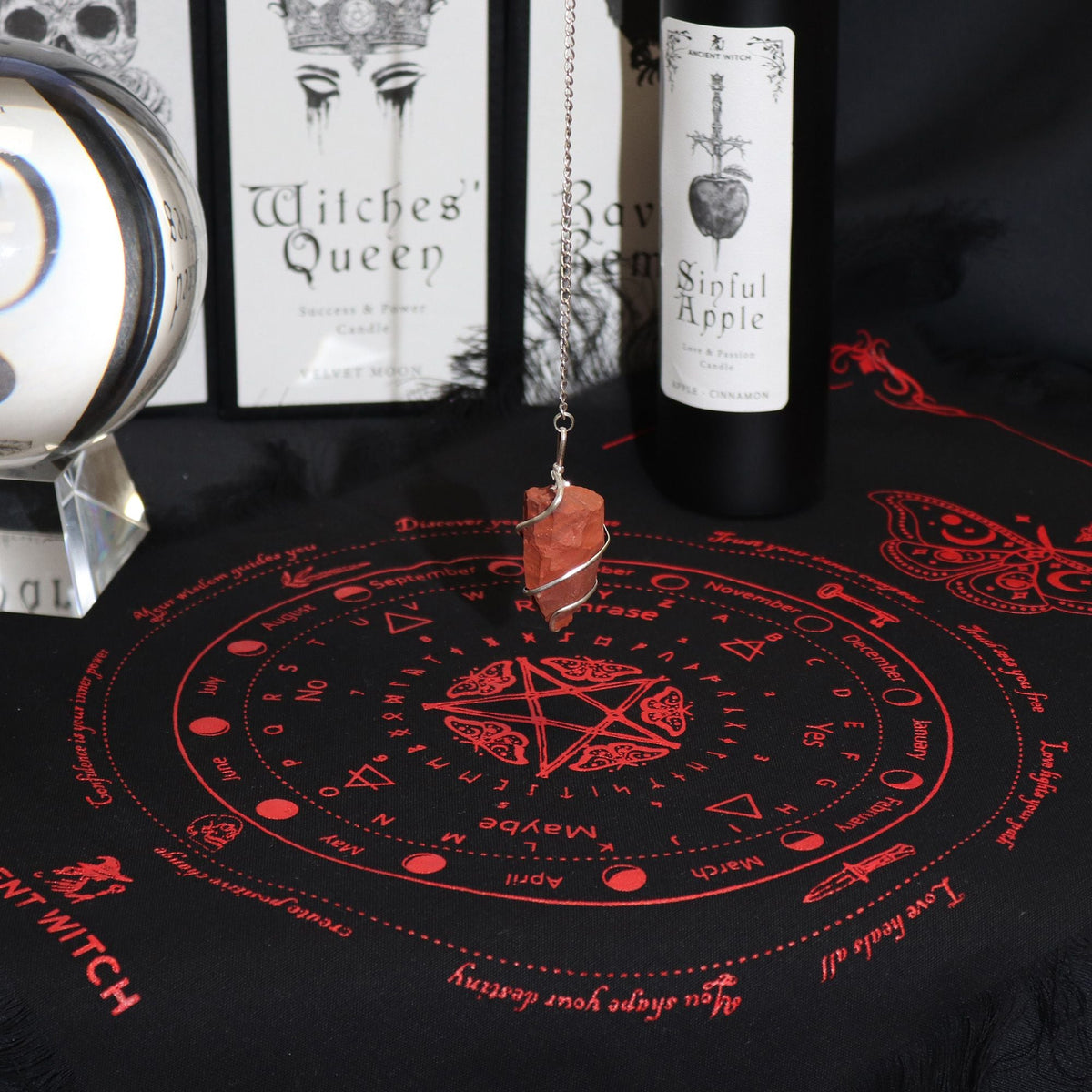 Red-and-black divination cloth with a hanging pendulum, shown with candles and packaging.
