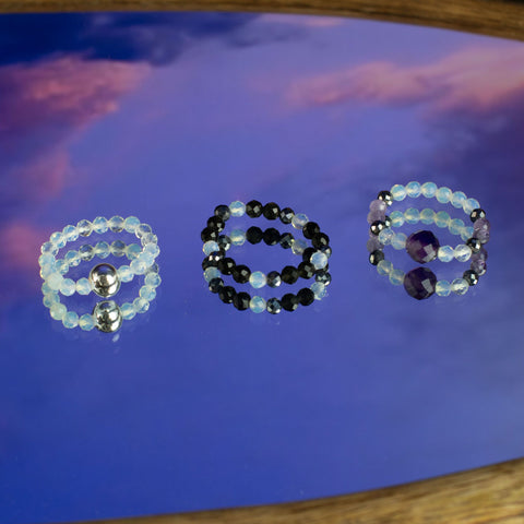 Three gemstone stretch rings in pale, black and amethyst tones, on a blue sky backdrop