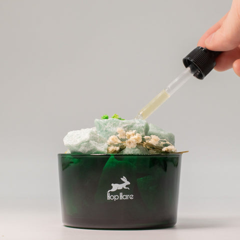 Green glass vessel with green aventurine chunks and dried flowers, a dropper adding oil from above