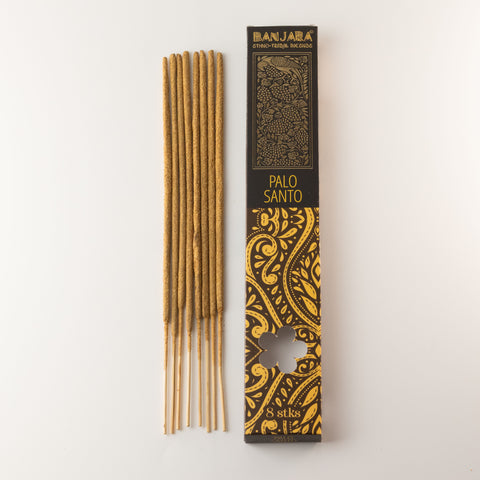 Long brown incense sticks arranged beside their black and yellow Palo Santo box on a pale background