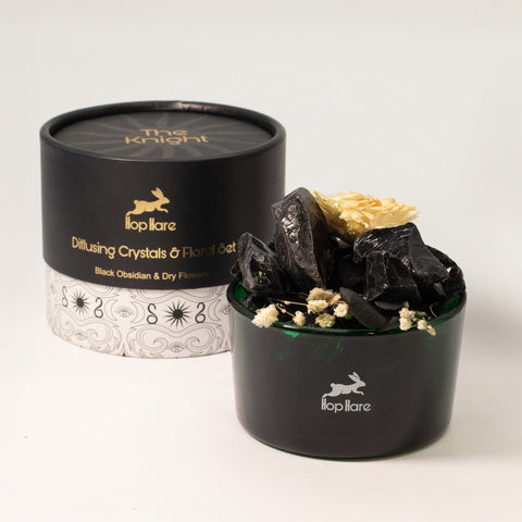 Black gift box with gold text beside a green jar of black obsidian chunks and a dried cream flower