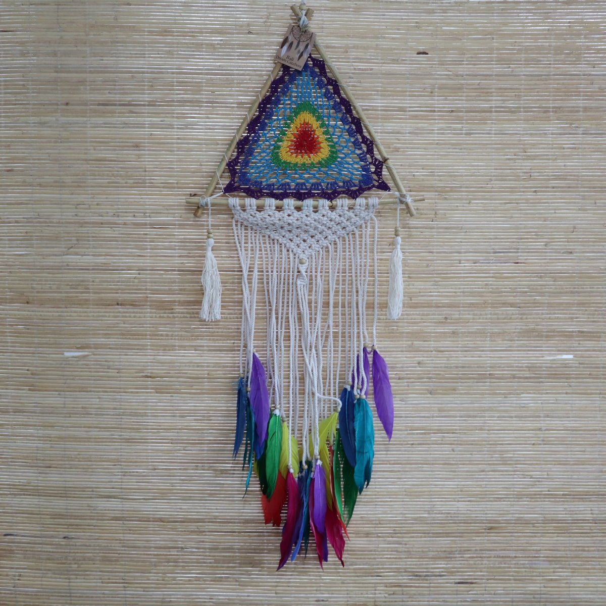 Multi-coloured pyramid dreamcatcher hanging against a woven reed screen, shown full length with white cords and feathers